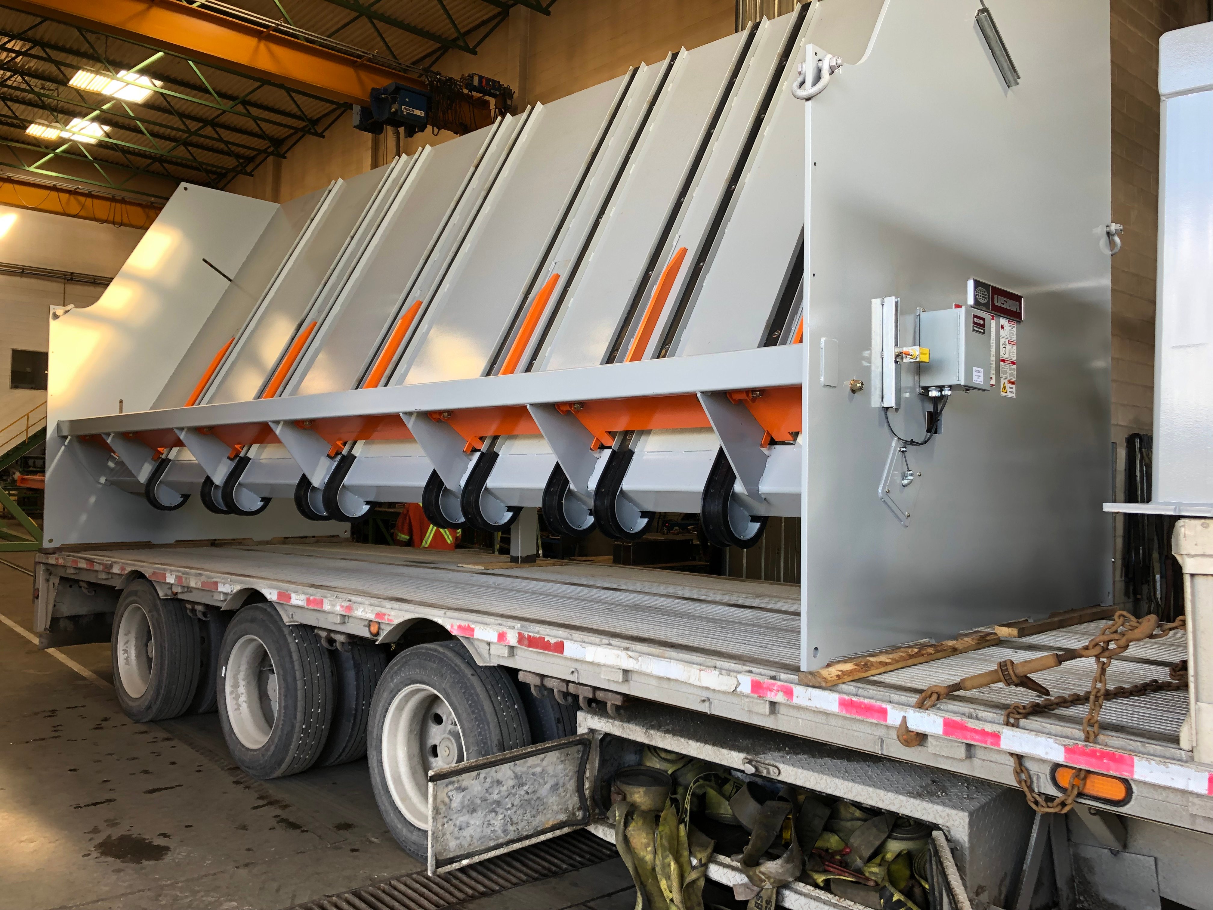 Industrial machinery loaded on flatbed trailer in a warehouse setting, featuring large metal construction with wheels and orange accents. Industrial machinery loaded on flatbed trailer in a warehouse setting, featuring large metal construction with wheels and orange accents.